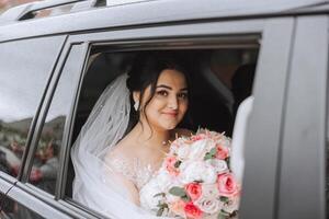 A beautiful bride, sitting in a car, looks out of the open window of the car. A beautiful bride with a bouquet of flowers in her hands is sitting in a stylish expensive car. photo