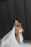 Beautiful young bride girl wearing a white dress with a long veil and posing near a gray wall in a modern city. photo