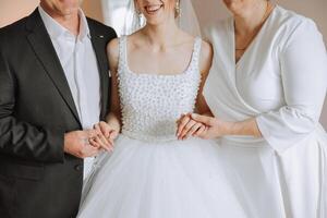 Happy parents hold the hands of the bride who is getting married and support her, wearing a wedding dress and a long veil. photo