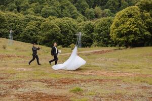 Photo of a young bride and groom walking together with a videographer on green grass against the background of mountains during a wedding video shoot, side view. Happy wedding couple, copy space.