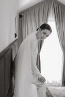 A beautiful and confident bride standing on a bed in white pajamas with a long veil in a hotel room poses for a photographer. Wedding concept. Morning of the bride. Preparation for the wedding. photo