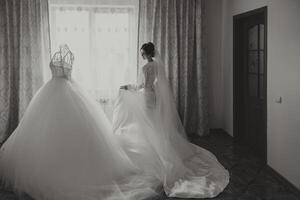 A beautiful brunette bride is getting ready for the wedding in a beautiful boudoir style outfit standing next to her wedding dress by the window. Black and white photo. photo