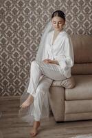 The bride in a silk suit and veil poses in her room, sitting on the couch. Morning of the bride. Preparation. Wedding photography photo