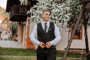 The groom in a black vest and black pants fastens a button and poses, against the background of a blooming tree. Wedding portrait. photo