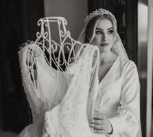 A bride in a silk suit with a veil poses in her room next to her wedding dress. Morning of the bride. Preparation. Black and white photo