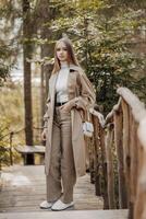 Vertical photo. Beautiful carefree young teenage girl in brown coat and white jeans and white sweater. Portrait of a beautiful girl in a brown raincoat against the background of the forest and nature. photo