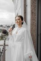 A beautiful bride with a long veil on her hair, wearing an elegant white peignoir, standing on a balcony overlooking the city, drinking a drink during the wedding morning. photo