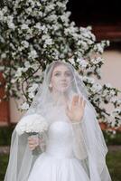 A red-haired bride poses against the background of a blooming tree. Magnificent dress with long sleeves, open bust. Spring wedding photo