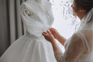 un contento novia es preparando para su lujoso Boda en un hotel habitación, con un Boda vestir en un maniquí cercano. retrato de un mujer con de moda cabello, maquillaje y un sonrisa en un vendaje vestido. foto