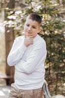 Close-up vertical portrait of a teenager in a white sweater and brown pants. Happy smiling teenager in summer park in sunlight. A beautiful child is looking at the camera in the clearing. photo