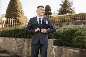 The groom in a black suit adjusts his jacket, poses against the background of a green tree. Wedding portrait. photo