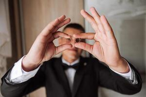 the groom holds the wedding rings with his fingers at the level of his eyes and looks into the camera through the rings. Funny photo. Selective focus. photo