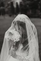 The bride in a luxurious dress, wrapped in a veil, sniffs her bouquet and poses for the camera. Black and white photo. Magnificent dress with long sleeves, open bust. Spring wedding photo