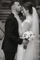 The groom gently hugs the bride and touches her forehead with his lips, the bride puts her hands on the groom's shoulders and enjoys the moment. Black and white photo