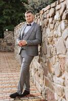 Groom in a gray suit, posing against the background of a stone wall. Wedding portrait. photo