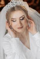 Wedding portrait. Blonde bride in a silk suit and tiara poses behind a sheer veil while sitting in a chair in her room photo