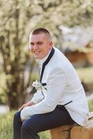The groom in a white jacket and black trousers poses while sitting on a wooden chair. Wedding portrait. photo