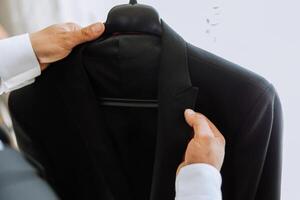 the man holds his black jacket on a hanger in his hands. The groom is preparing for the wedding ceremony. Detailed close-up photo of hands