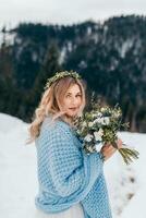 vertical retrato de un linda novia con un ramo de flores de flores en su manos, envuelto en un azul frazada, esperando para el novio en contra el antecedentes de un invierno paisaje. foto