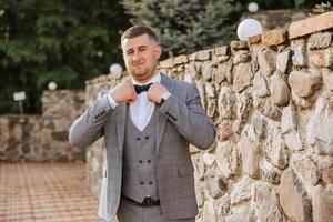The groom in a gray suit adjusts his bow tie, poses against the background of a stone wall. Wedding portrait. photo