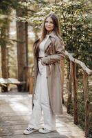 Vertical photo. Beautiful carefree young teenage girl in brown coat and white jeans and white sweater. Portrait of a beautiful girl in a brown raincoat against the background of the forest and nature. photo