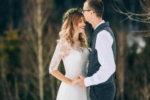 Portrait of the bride and groom against the background of a pine forest. The bride in a white wedding dress with a wreath on her head, the groom in a white shirt and vest. Winter wedding concept. photo