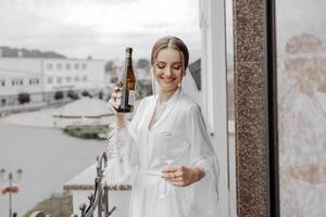 A beautiful bride with a long veil on her hair, wearing an elegant white peignoir, standing on a balcony overlooking the city, drinking a drink during the wedding morning. photo