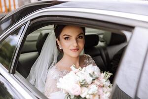 A beautiful bride, sitting in a car, looks out of the open window of the car. A beautiful bride with a bouquet of flowers in her hands is sitting in a stylish expensive car. photo