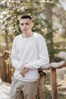 Close-up vertical portrait of a teenager in a white sweater and brown pants. Happy smiling teenager in summer park in sunlight. A beautiful child is looking at the camera in the clearing. photo