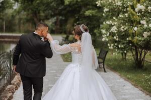 A beautiful young bride is walking with her groom in the summer park. Photo of the bride and groom from behind. Beautiful wedding white dress. Walks in the park. A happy and loving couple.