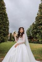 Curly brunette bride in a lush veil and dress with long sleeves, holding a bouquet and posing against a background of green trees. Spring wedding photo