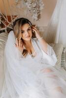 Morning of the bride before the wedding. A beautiful young woman with a veil and a beautiful hairstyle in a white robe in a room with a wonderful interior. Natural beauty and professional makeup. photo