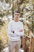 Close-up vertical portrait of a teenager in a white sweater and brown pants. Happy smiling teenager in summer park in sunlight. A beautiful child is looking at the camera in the clearing. photo