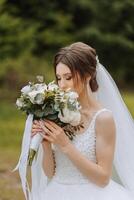 The bride in a lush dress and veil, holding a bouquet of white flowers and greenery, posing against the background of green trees.. Spring wedding photo