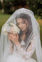 el novia en un lujoso vestido, envuelto en un velo, olfatea su ramo de flores y poses para el cámara. magnífico vestir con largo mangas, abierto busto. primavera Boda foto