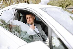 a handsome groom sits with a wedding bouquet inside a white car and looks out the window. A happy moment on the wedding day. photo