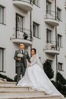 A bride in a white dress with a train and a groom in a suit pose on the steps of a building. Wedding photo session in nature