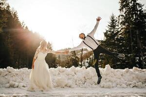 The bride and groom are running along a snowy road against the background of a pine forest and beautiful contrast sunlight. Side view. Winter wedding. Place for logo. photo