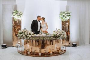 A young groom in a black suit and a smiling blonde bride at their table in a banquet hall, looking at each other and kissing against a background of fresh flowers and candles in glass bulbs. photo