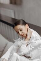 hermosa y confidente novia sentado en cama en blanco pijama con largo velo en hotel habitación. moderno Corte de pelo y constituir. Boda concepto. Mañana de el novia. preparación para el Boda ceremonia foto