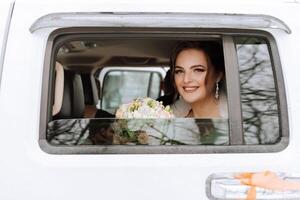 A beautiful bride with a bouquet of flowers is looking at the camera while sitting in a stylish white car. photo