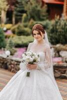 A red-haired bride twirls her long veil against a background of wooden houses. Magnificent dress with long sleeves, open bust. Summer wedding photo