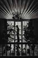 A stylish groom is standing in front of the window and preparing for the wedding ceremony. Groom's morning. Businessman wears a shirt. Black and white photo in profile.