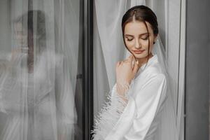 retrato de novia niña en blanco vestido con profesional peinado y natural maquillaje en hotel habitación con reflexión en ventana. el mejor día foto