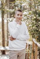 Close-up vertical portrait of a teenager in a white sweater and brown pants. Happy smiling teenager in summer park in sunlight. A beautiful child is looking at the camera in the clearing. photo