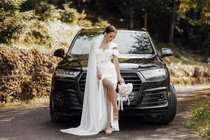 A dark-haired bride stands next to a modern black car on your wedding day with a bouquet of flowers. Portrait of the bride. White dress, long veil photo