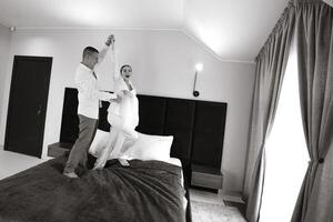 The morning of the bride and groom in a hotel room. Happy and in love bride and groom jumping and having fun on the bed. Confident girl-bride and handsome groom. Preparation for the wedding photo