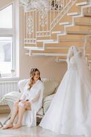 Morning of the bride before the wedding. A beautiful young woman with a veil and a beautiful hairstyle in a white robe near her wedding dress on a mannequin. Professional makeup. Last preparations. photo