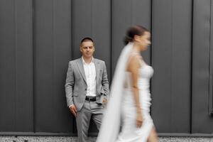 Fashionable and staged photo of the bride and groom against the background of a gray wall. A bride with a long veil blowing in the wind. The bridegroom is looking at the camera