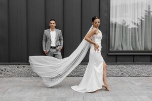 Fashionable and staged photo of the bride and groom against the background of a gray wall. A bride with a long veil blowing in the wind. The bridegroom is looking at the camera